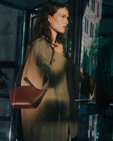 Person in oversized coat holding a shoulder bag, standing near glass doors.