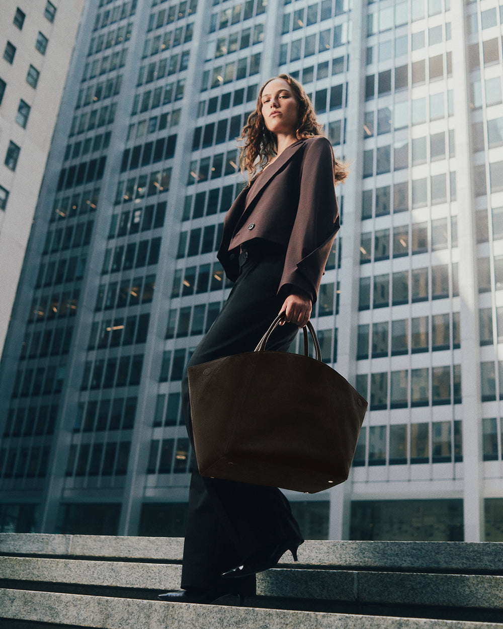 A woman standing outdoors holding a large bag with tall buildings in the background.