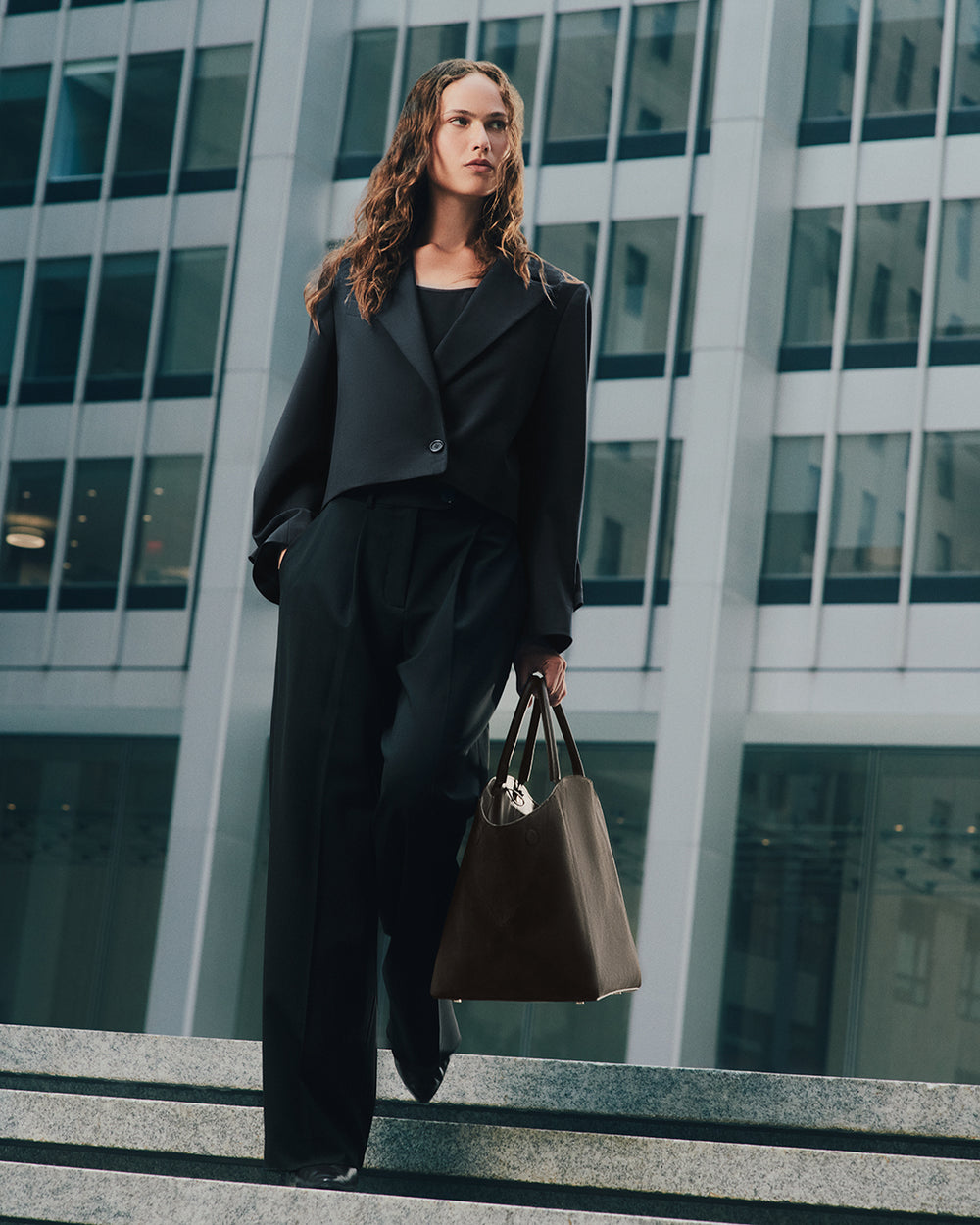 Person in suit holding a large handbag, standing on steps near tall buildings.