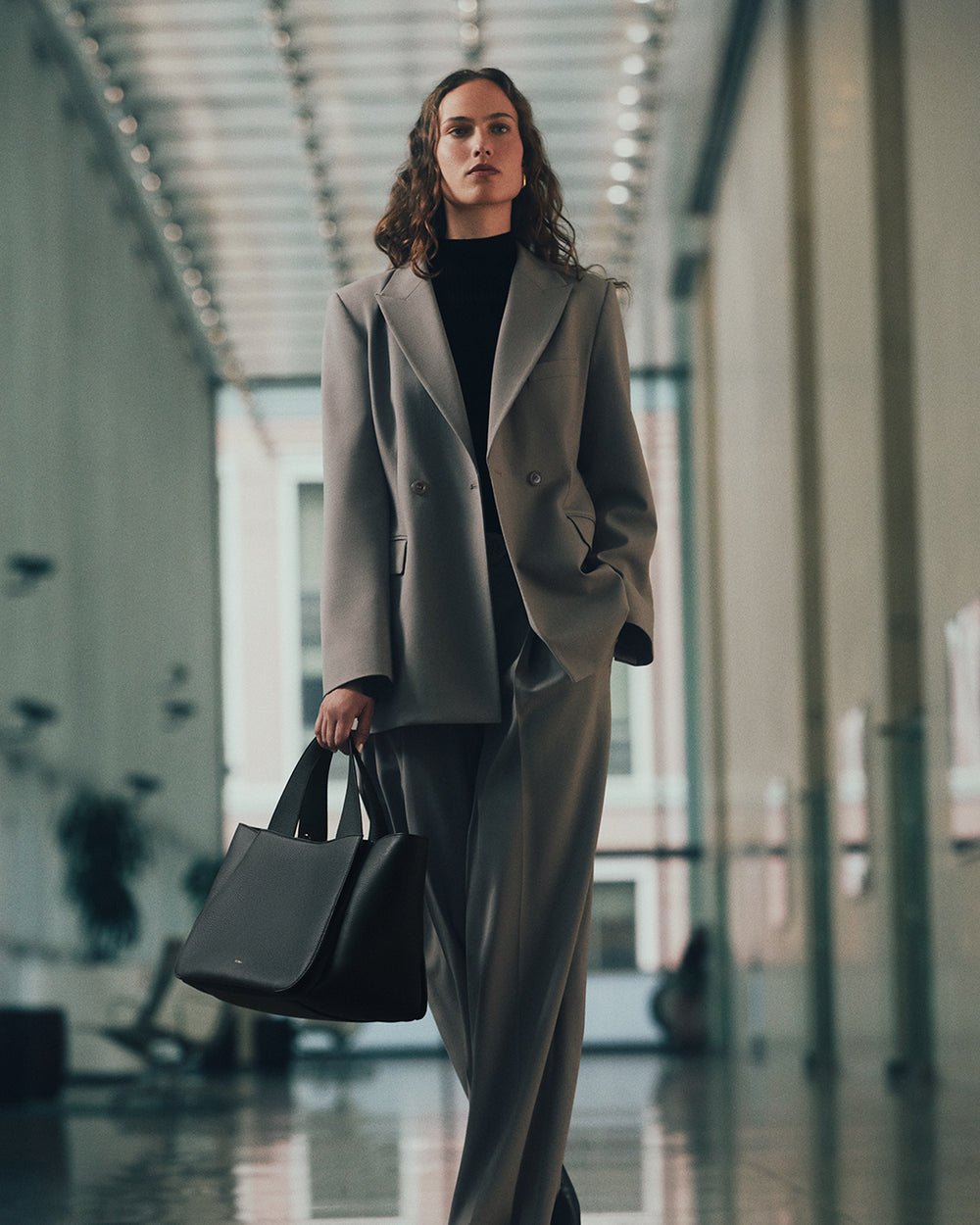 Person in suit holds a handbag walking in a corridor with large windows.