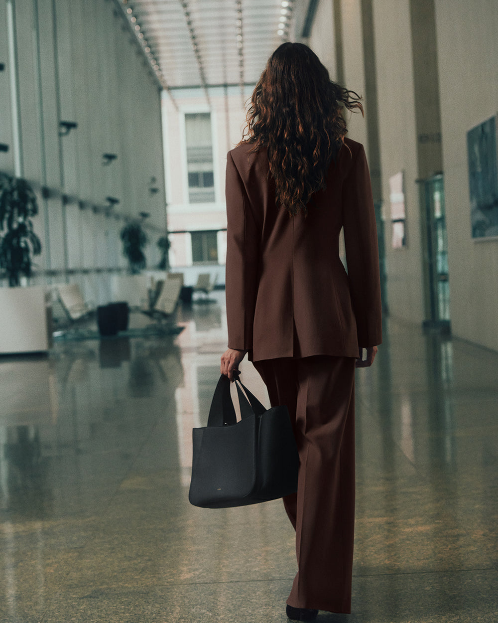 Person in a suit walking in a modern building, carrying a large handbag.
