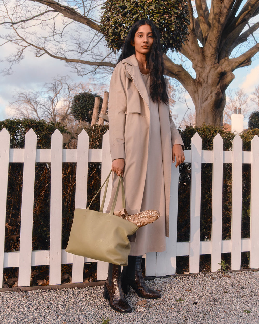 Woman standing by a white fence with a bag, near a tree.