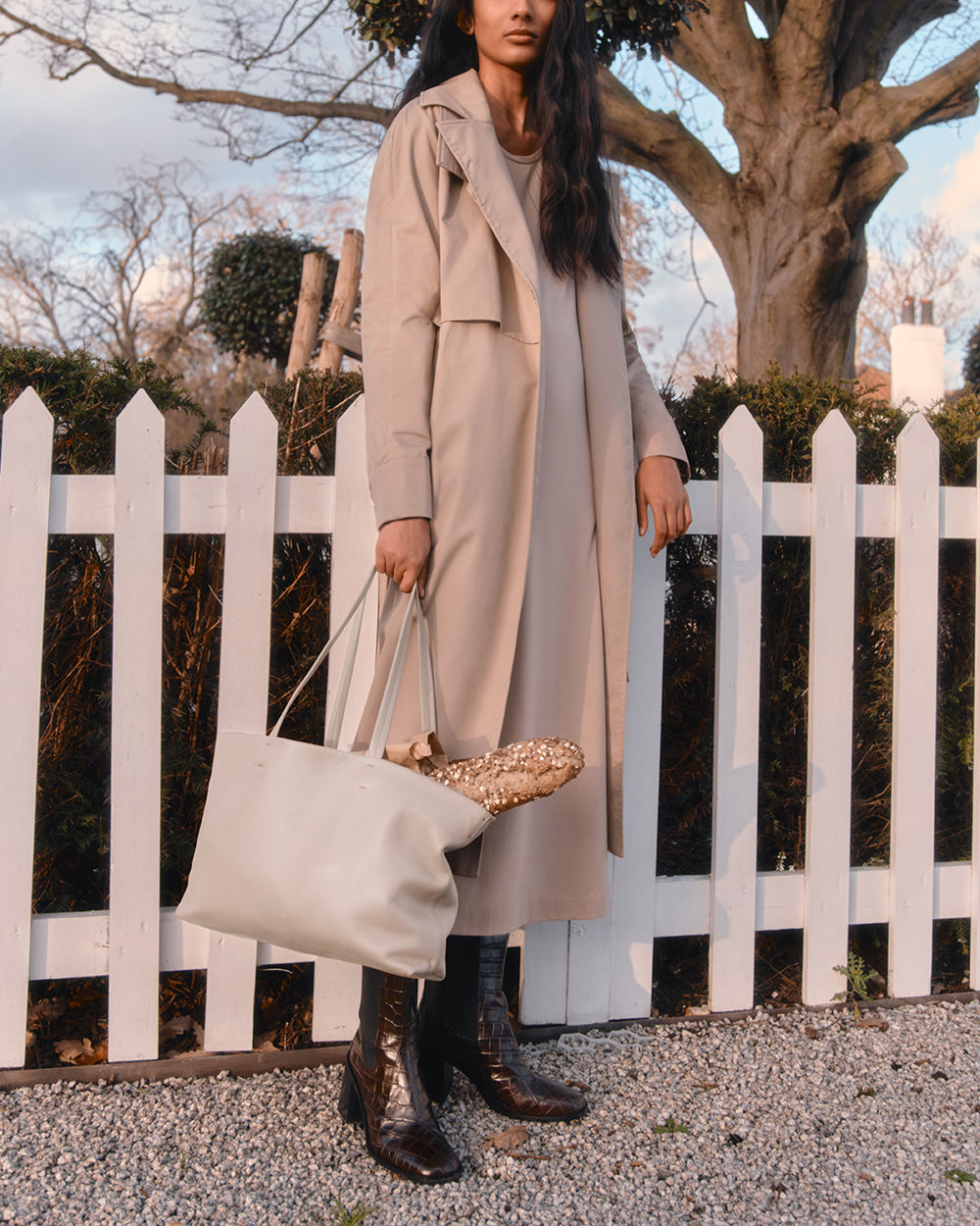 Person in long coat holding a tote bag stands by a white fence.
