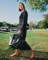 Person walking in park holding large tote bag, wearing a denim and sandals.