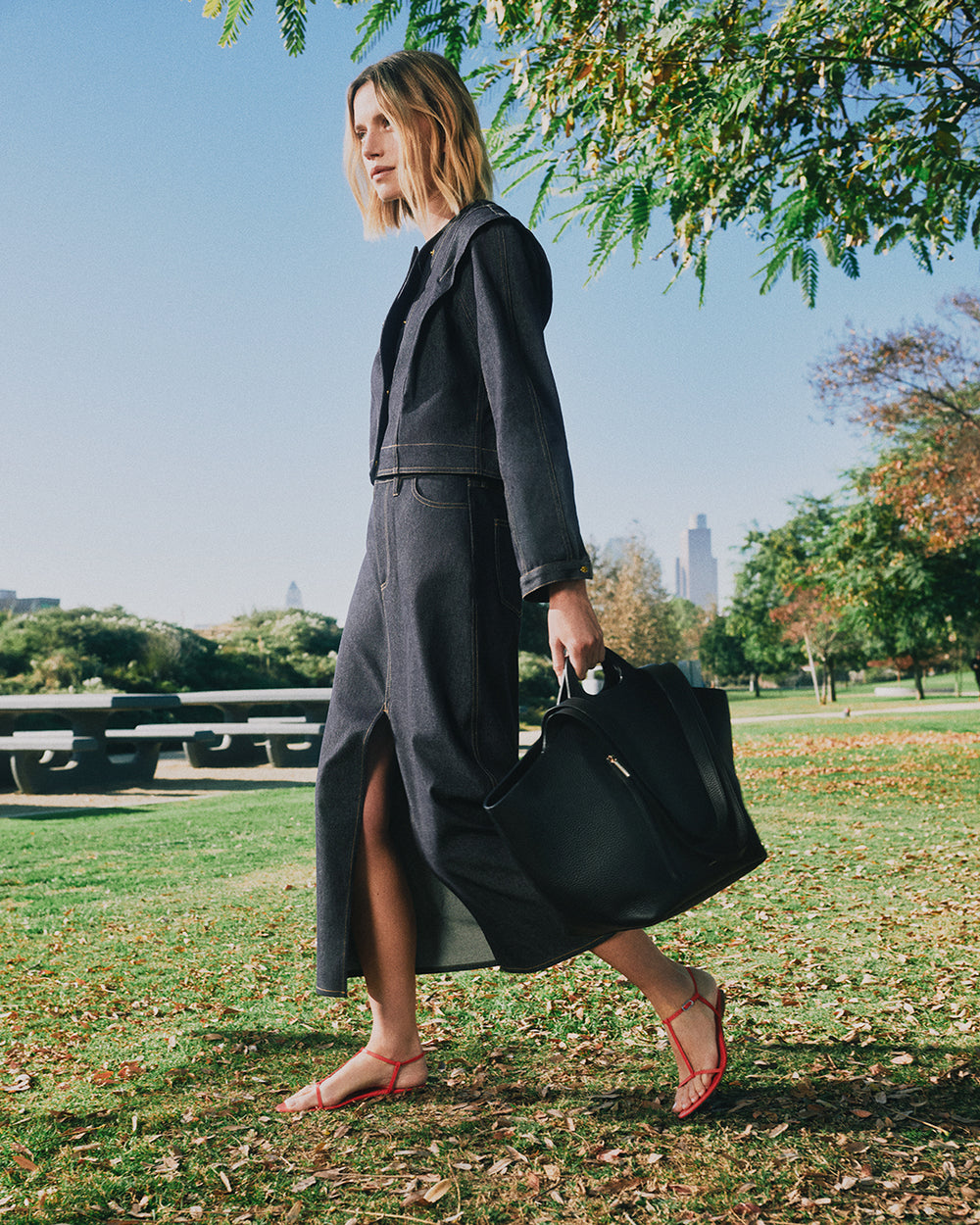 Person in a denim jacket, wearing a long skirt, holding a tote bag, walking in a park.