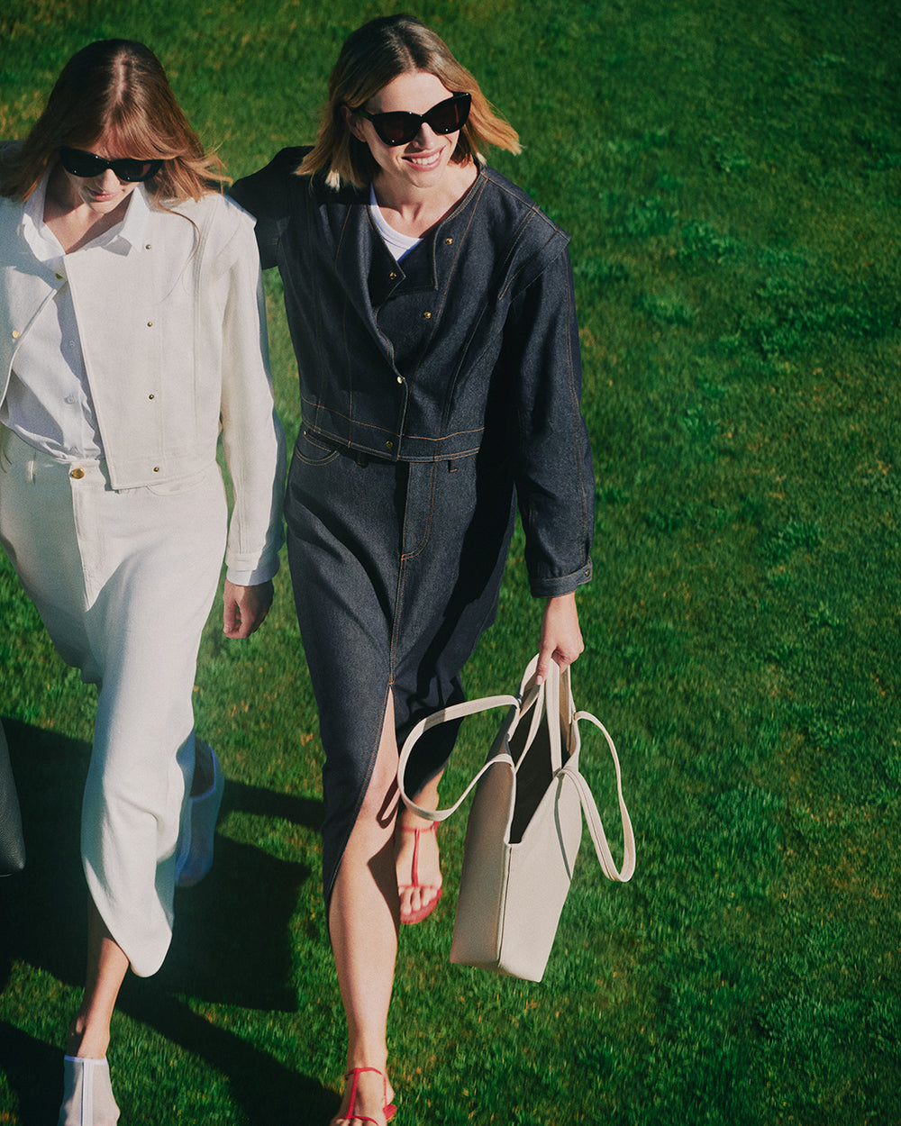 Two people walking on grass, wearing denim jackets and skirts, each holding a bag.