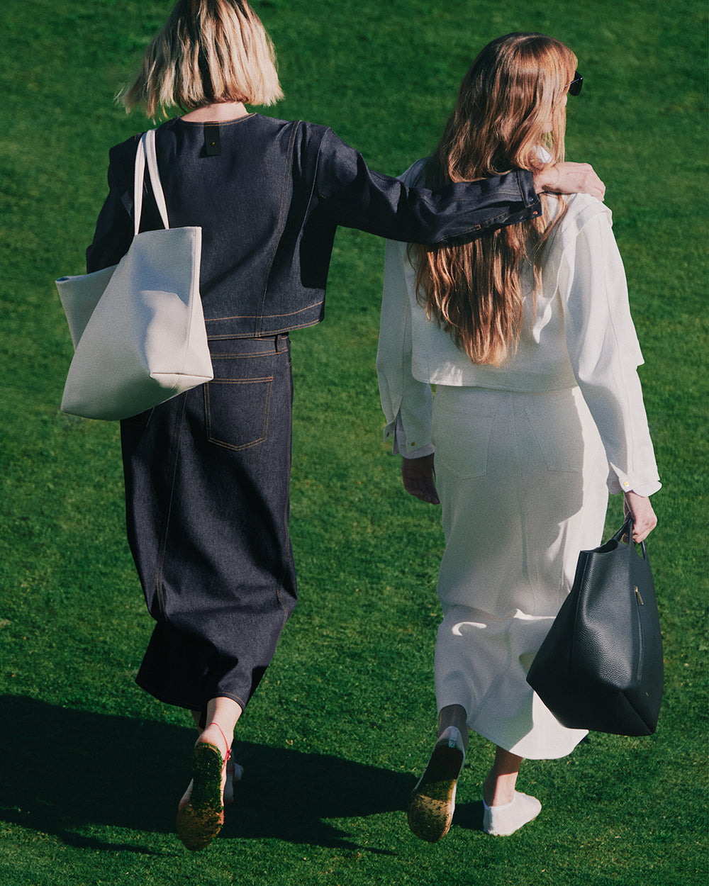 Two people walking on grass, wearing denim jackets and skirts, each holding a bag.