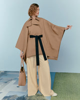 Woman standing, holding out cape, with a handbag and wide leg trousers.
