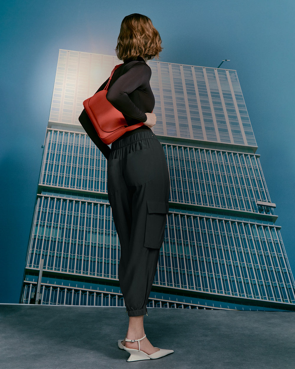 Woman facing a large building, carrying a backpack, viewed from the side.