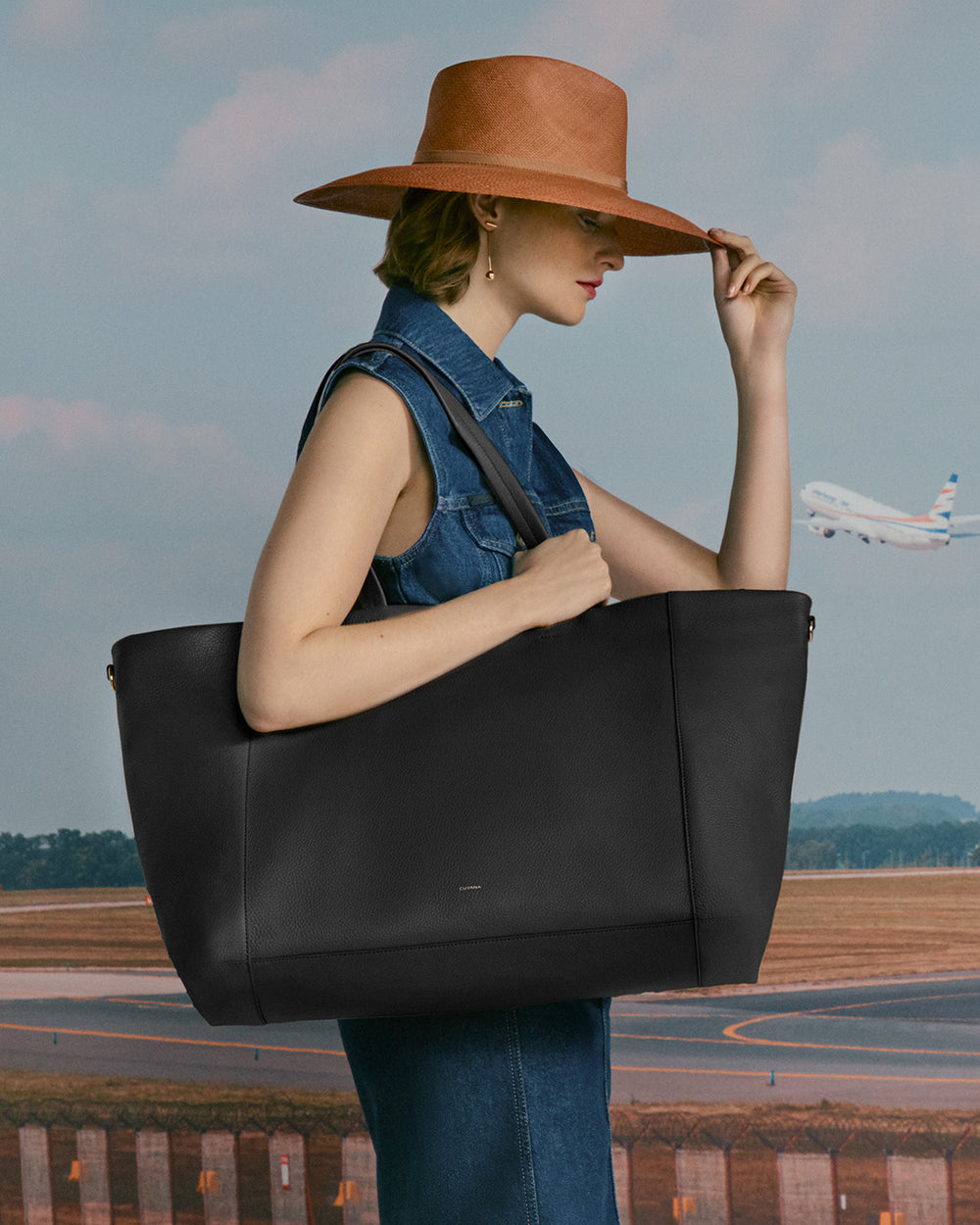 Person in a sleeveless outfit holding a large tote bag with a hat on, plane in the background.