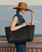 Woman holding a large bag and touching her hat at an airport with a plane in the background.