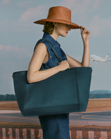 Person with hat holding a large tote bag at an airport with plane in background