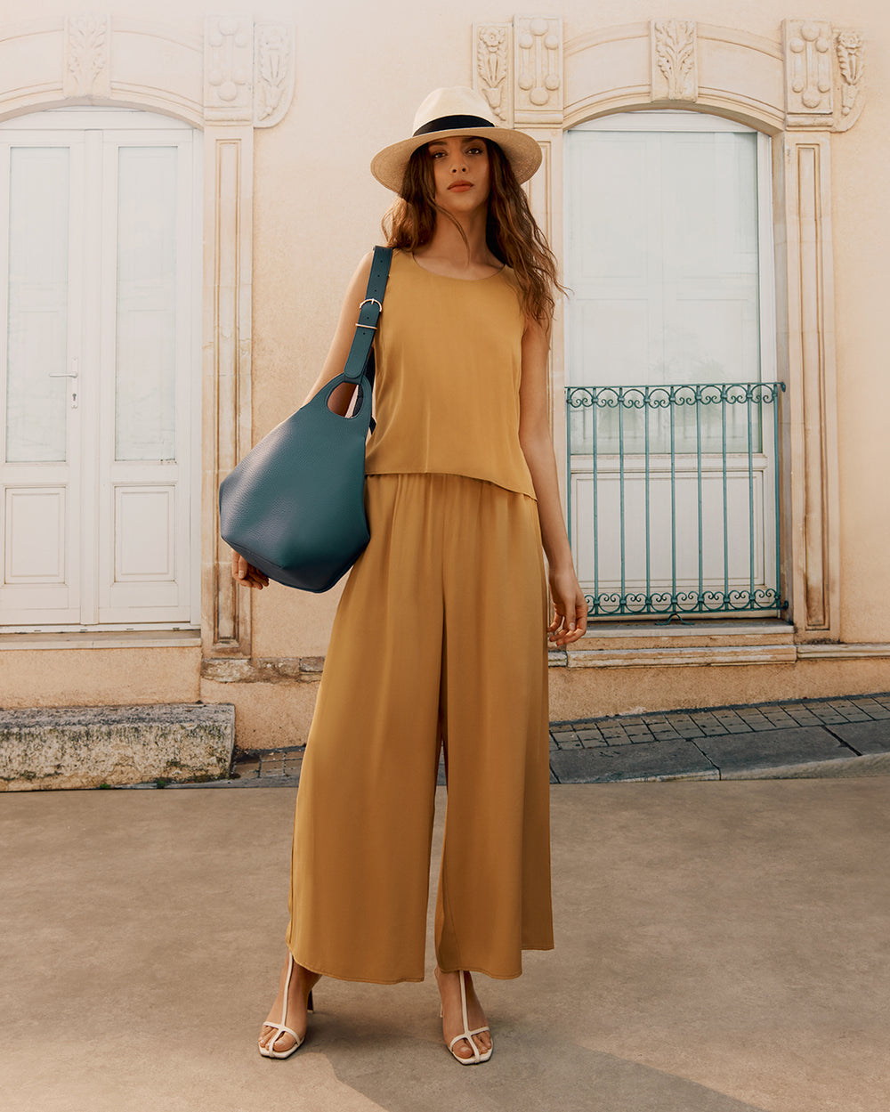 Woman standing in front of a building wearing a sleeveless outfit, hat, and holding a large bag