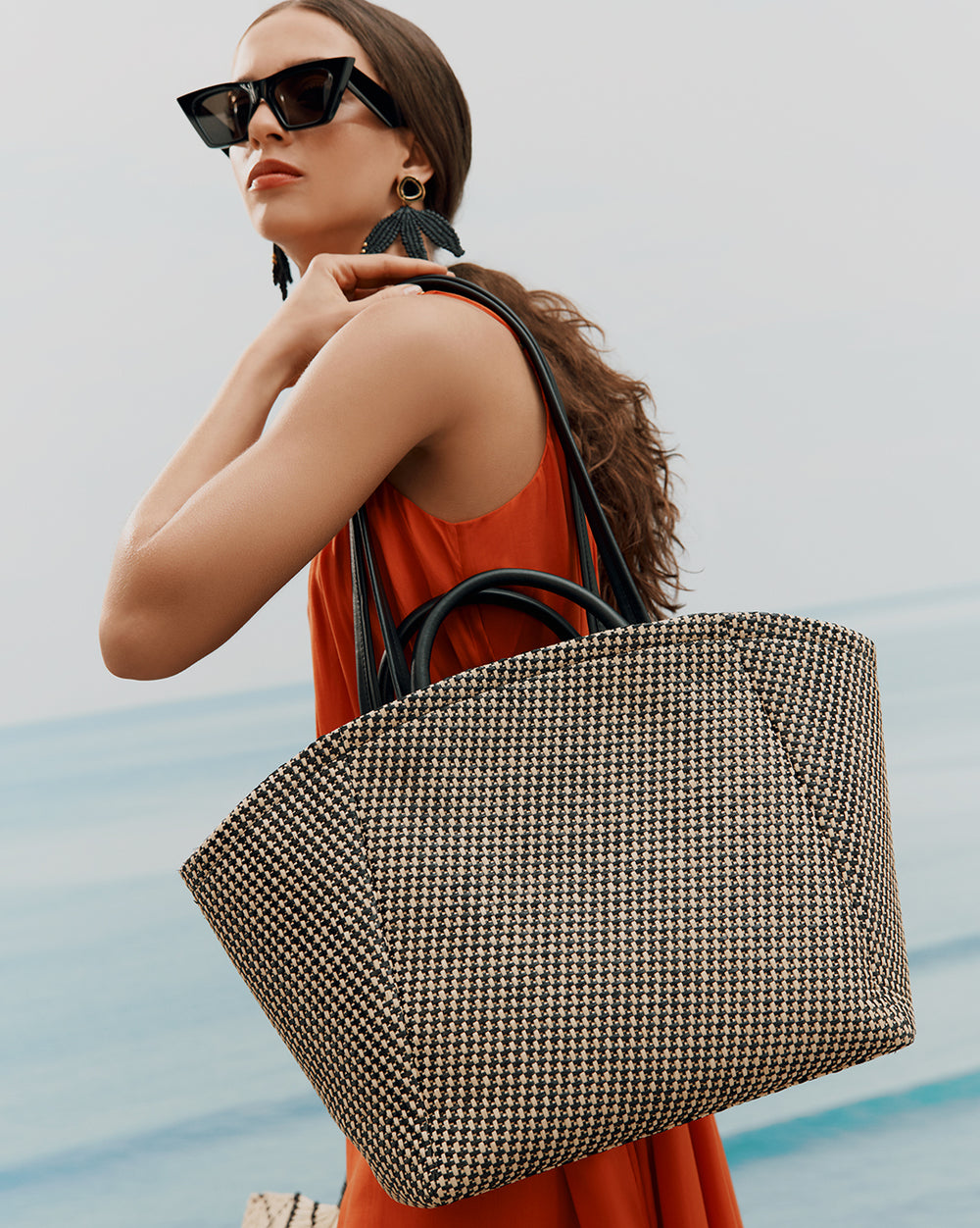 Woman holding a large tote bag, wearing sunglasses and earrings, looking away.