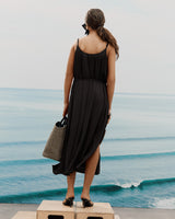 Person standing on platform by ocean, wearing long dress and carrying a woven structured handbag