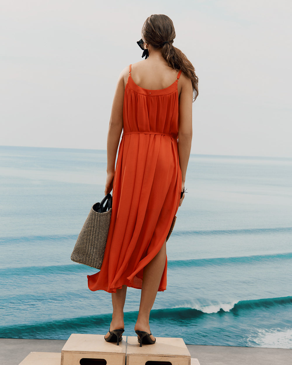 Woman in a dress standing on wood blocks, holding a woven tote, with an ocean backdrop.