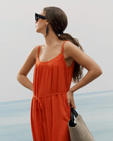 Person in dress wearing sunglasses, holding a travel bag, standing near the water looking to the side.