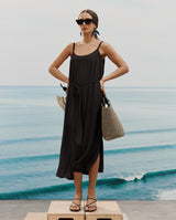 Woman in a dress standing by the sea with sunglasses and a bag.