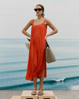 Person in a dress stands on a platform by the sea holding a bag in one hand