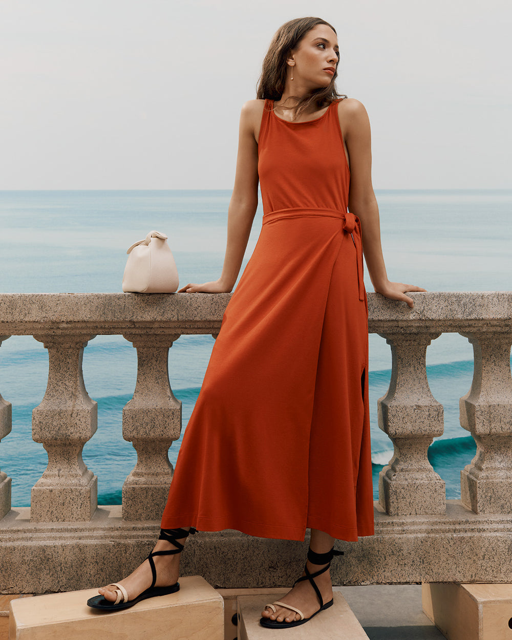 Woman in dress leaning on a stone rail near the sea; small bag on the rail.