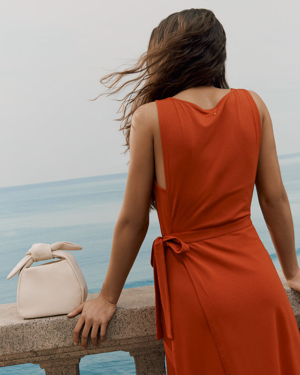 Person in a dress looking out over the ocean, with a small handbag on the stone railing.
