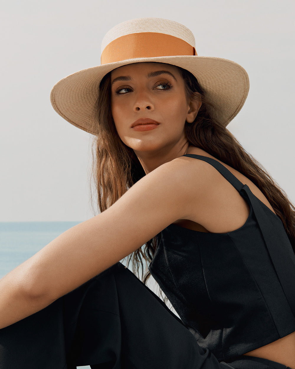 Woman in a hat sitting by the sea looking over her shoulder.