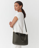 Woman standing sideways, holding a shoulder bag, looking over her shoulder.