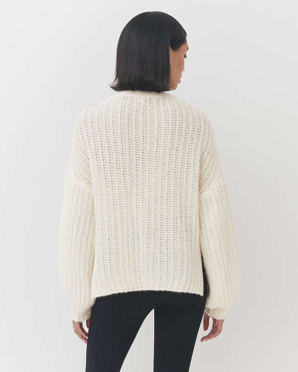 Woman in a sweater viewed from the back standing against a plain background.