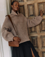 Person wearing a sweater and skirt with a shoulder bag standing in front of a wooden door.