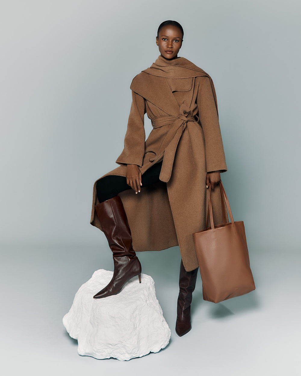 Person in stylish outfit with a coat, boots, and a bag posing with one foot on a rock.
