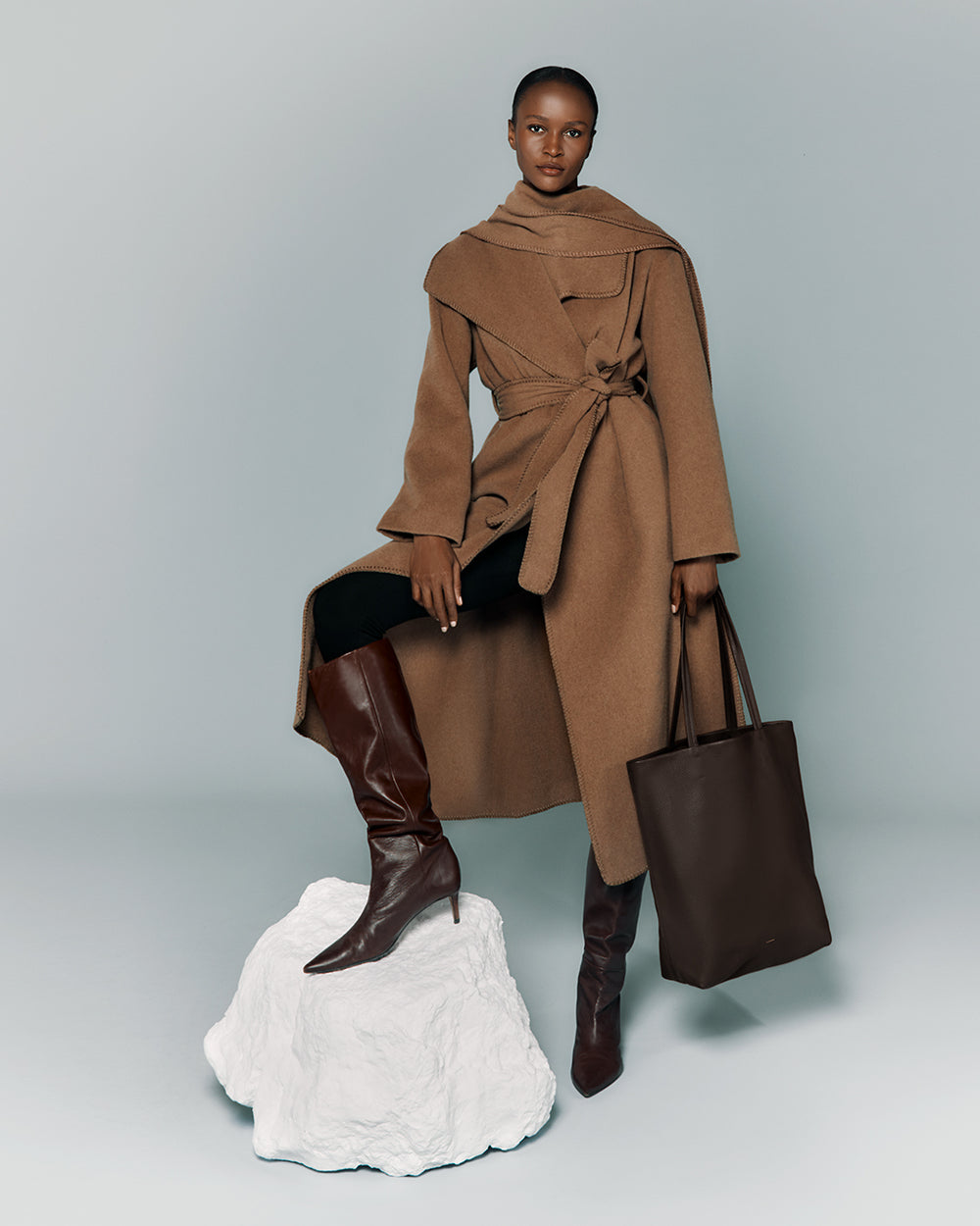 Person in coat with cape, holding a tote bag, one foot on a white platform, wearing tall boots.