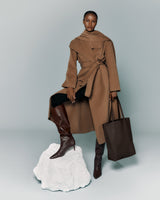 Person in coat with cape, holding a tote bag, one foot on a white platform, wearing tall boots.