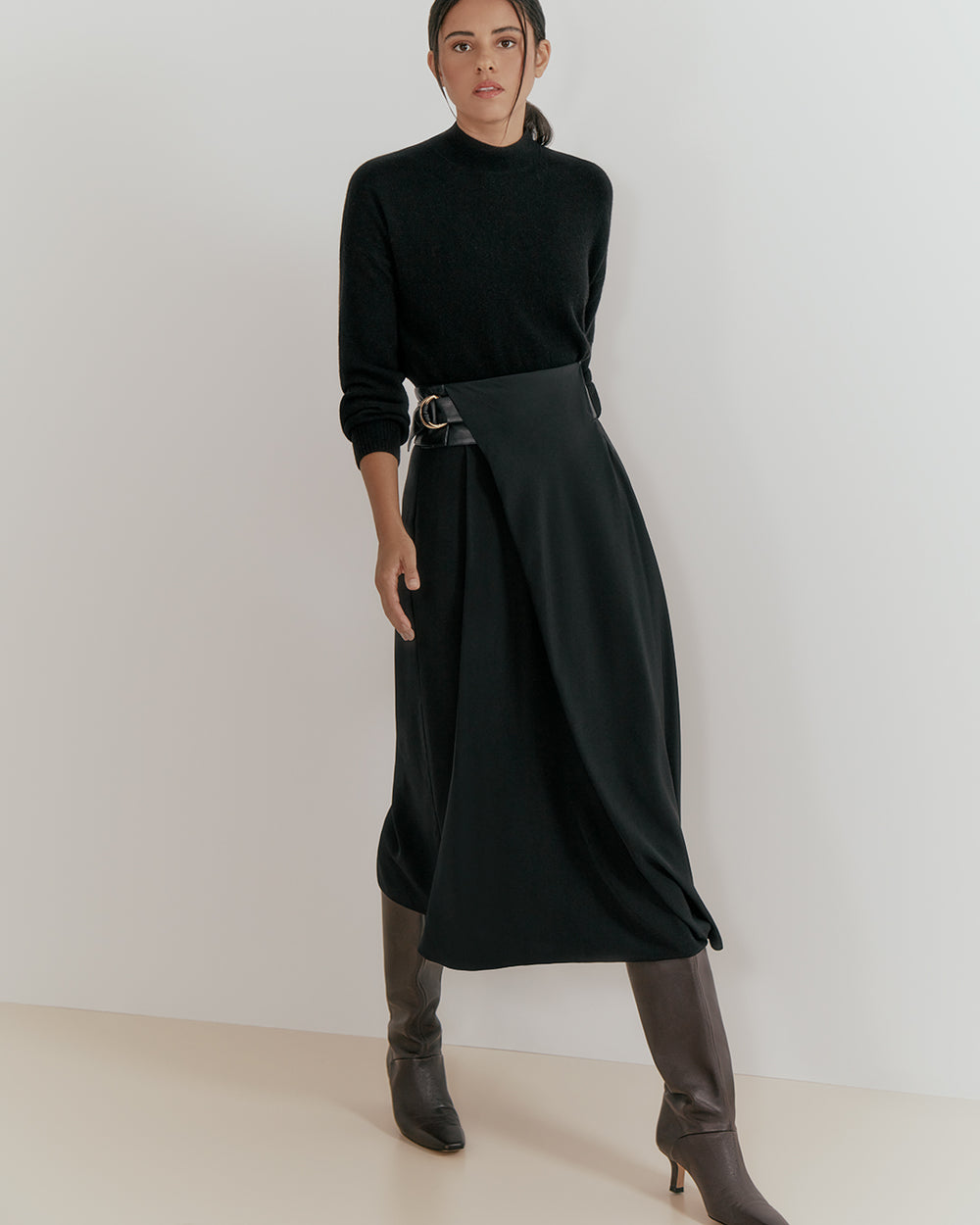 Woman standing in a turtleneck and belted skirt with boots.