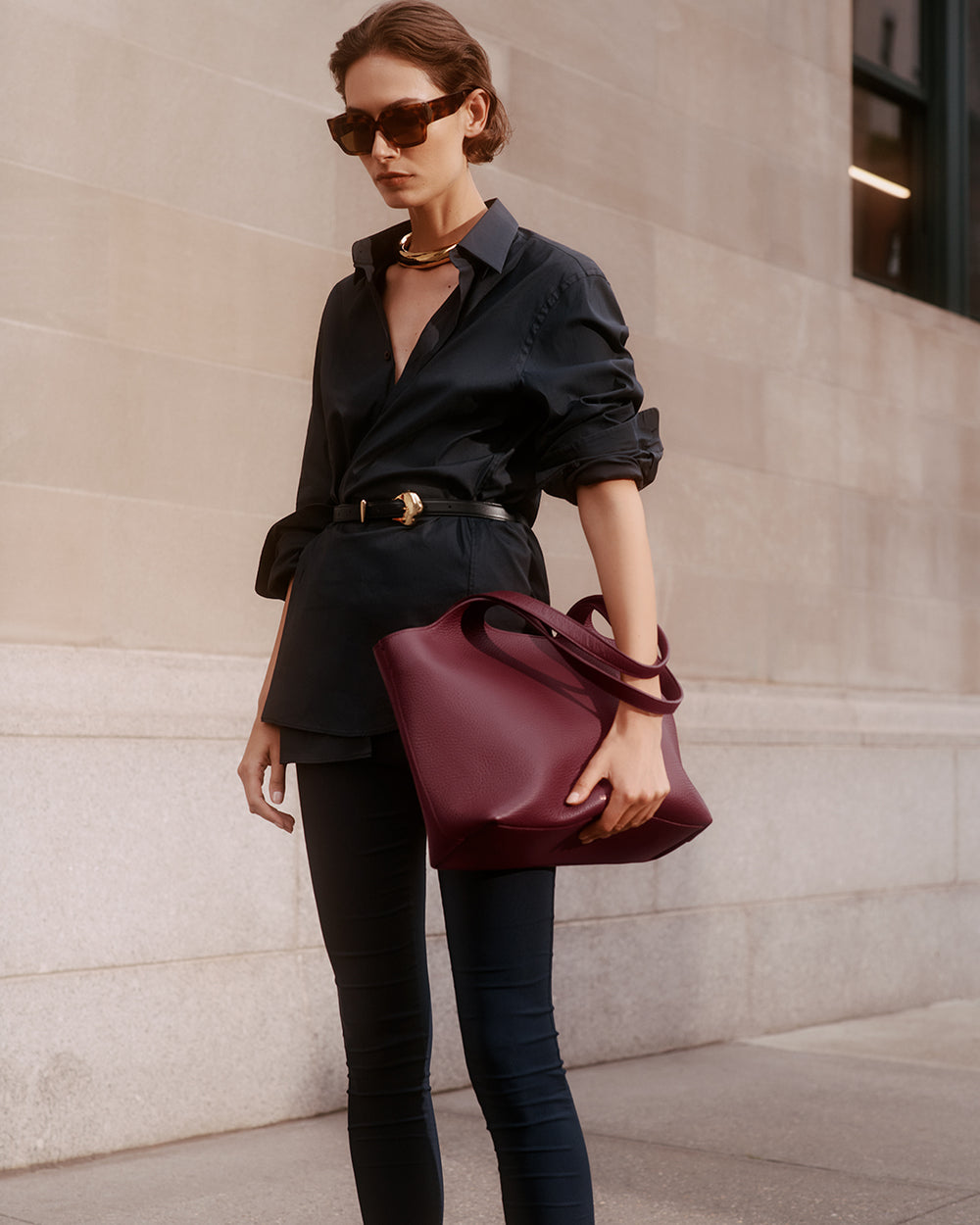 Person wearing dark outfit, sunglasses, holding a handbag, standing near a building