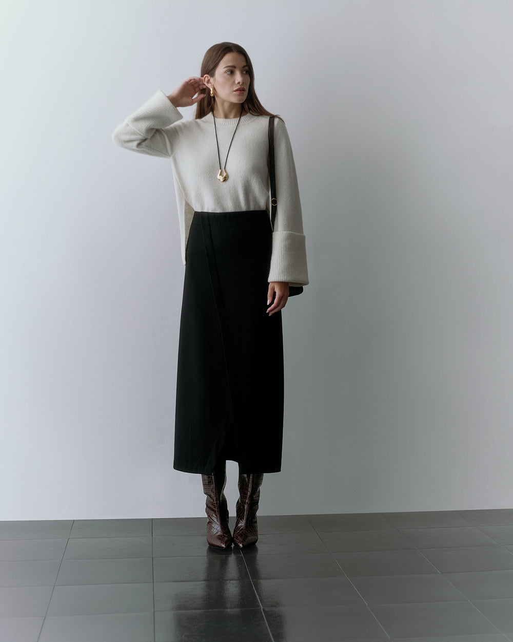 Woman wearing a long-sleeve shirt, long skirt, boots, and a pendant necklace standing indoors.