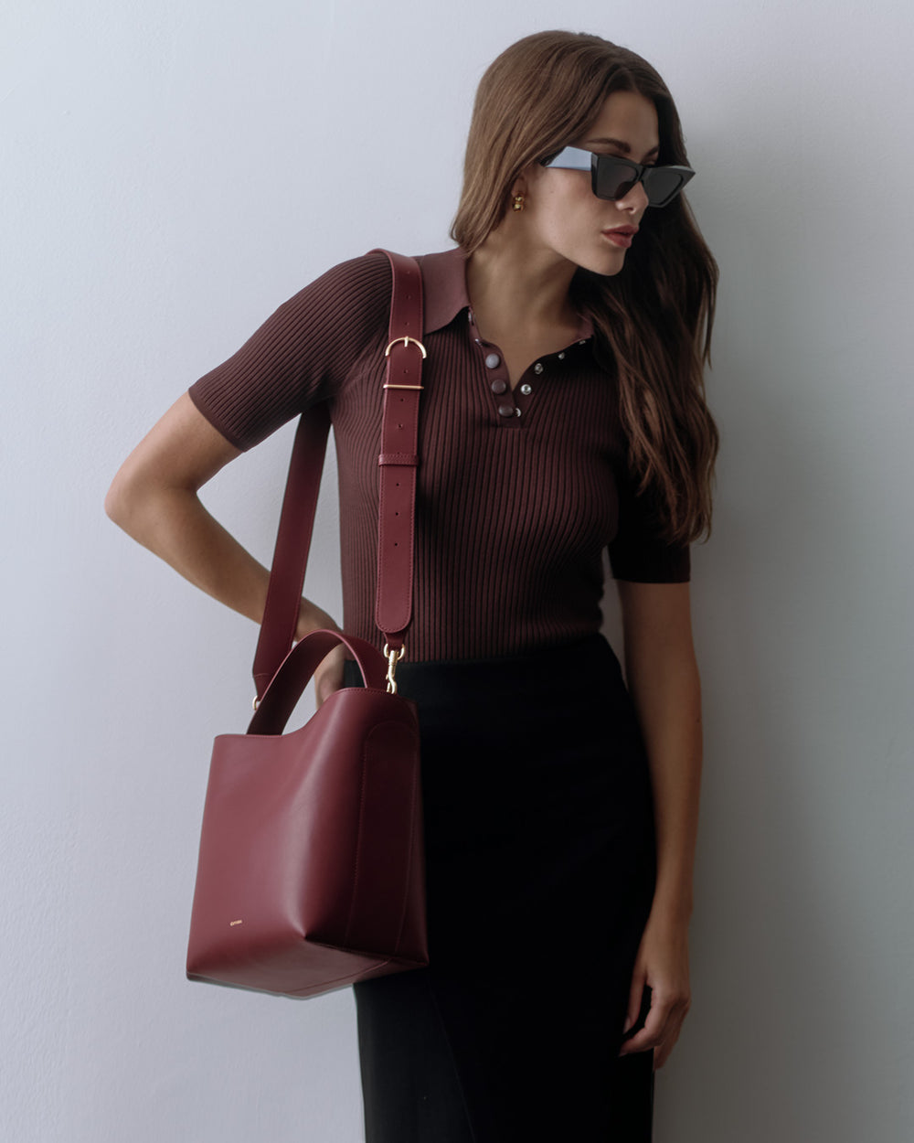 Person wearing sunglasses and carrying a large handbag over one shoulder standing against a wall