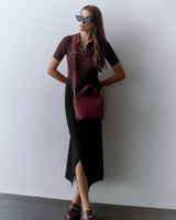 Woman wearing a fitted shirt, long skirt, sunglasses, and a shoulder bag, standing against a plain wall.