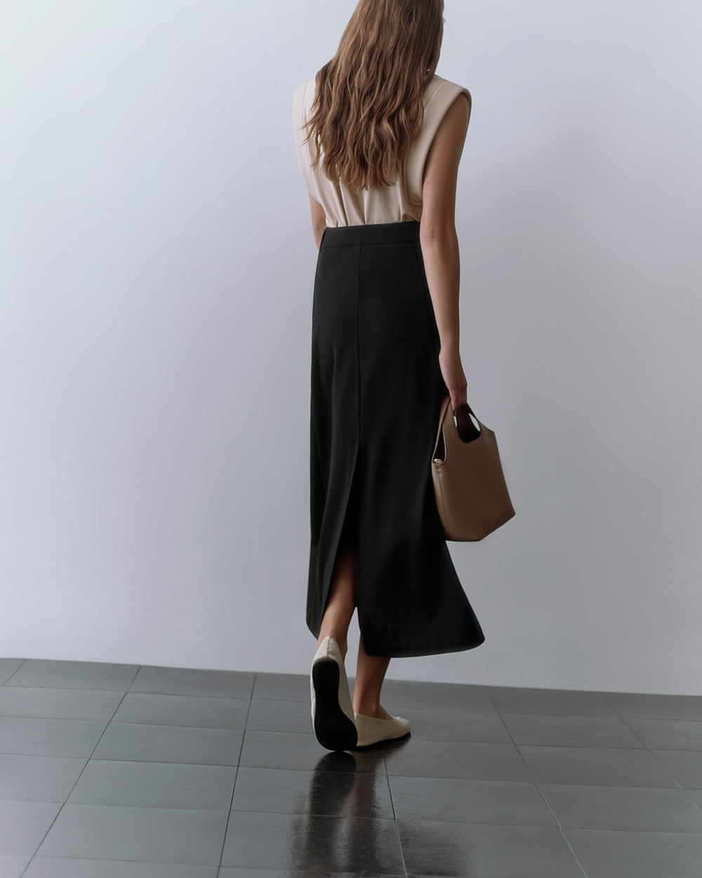 Person wearing sleeveless top and long skirt, holding a handbag, walking on tiled floor.