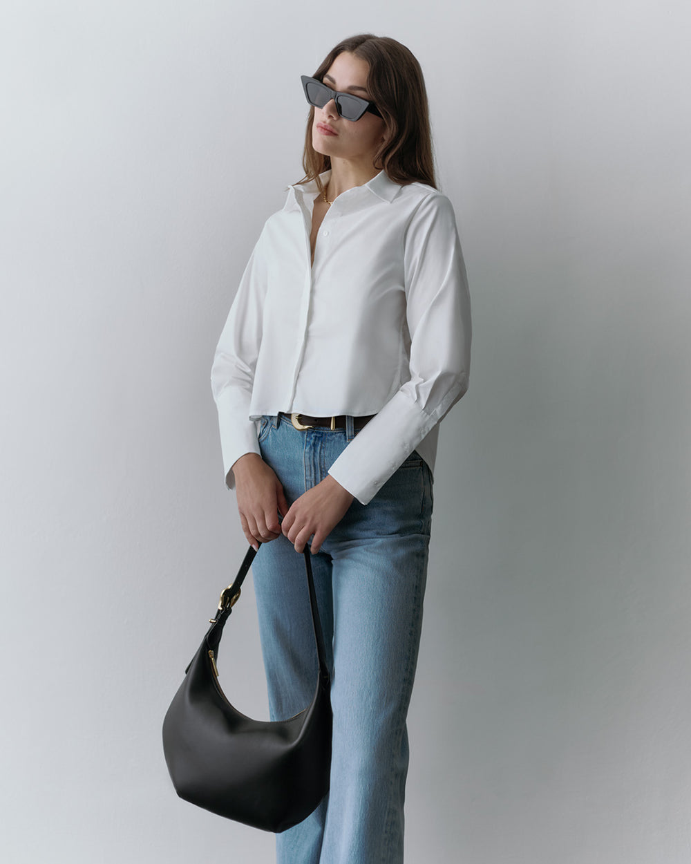 Woman wearing sunglasses, collared shirt, and jeans, holding a curved handbag in front of a plain background