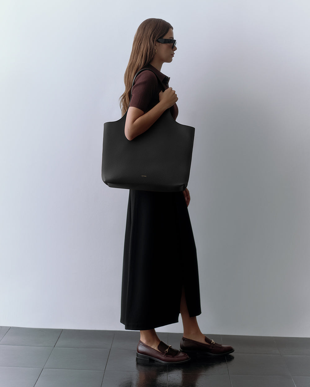 Woman wearing sunglasses, carrying large shoulder bag, in long skirt and loafers.