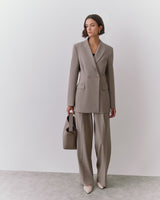Person in suit holding handbag, standing against plain backdrop.