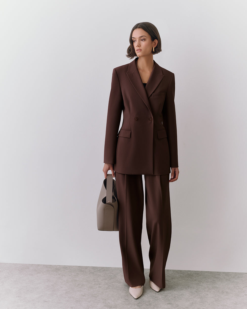 Person in a suit holding a handbag stands against a plain background.