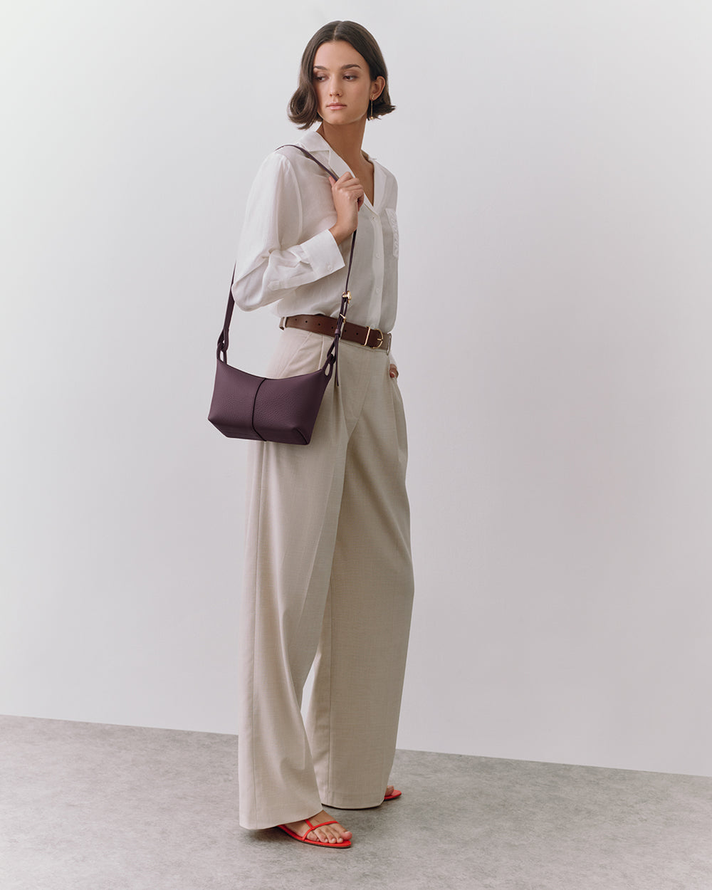 Person in formal outfit holding a shoulder bag, standing in a neutral space.