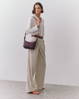 Person in formal outfit holding a shoulder bag, standing in a neutral space.