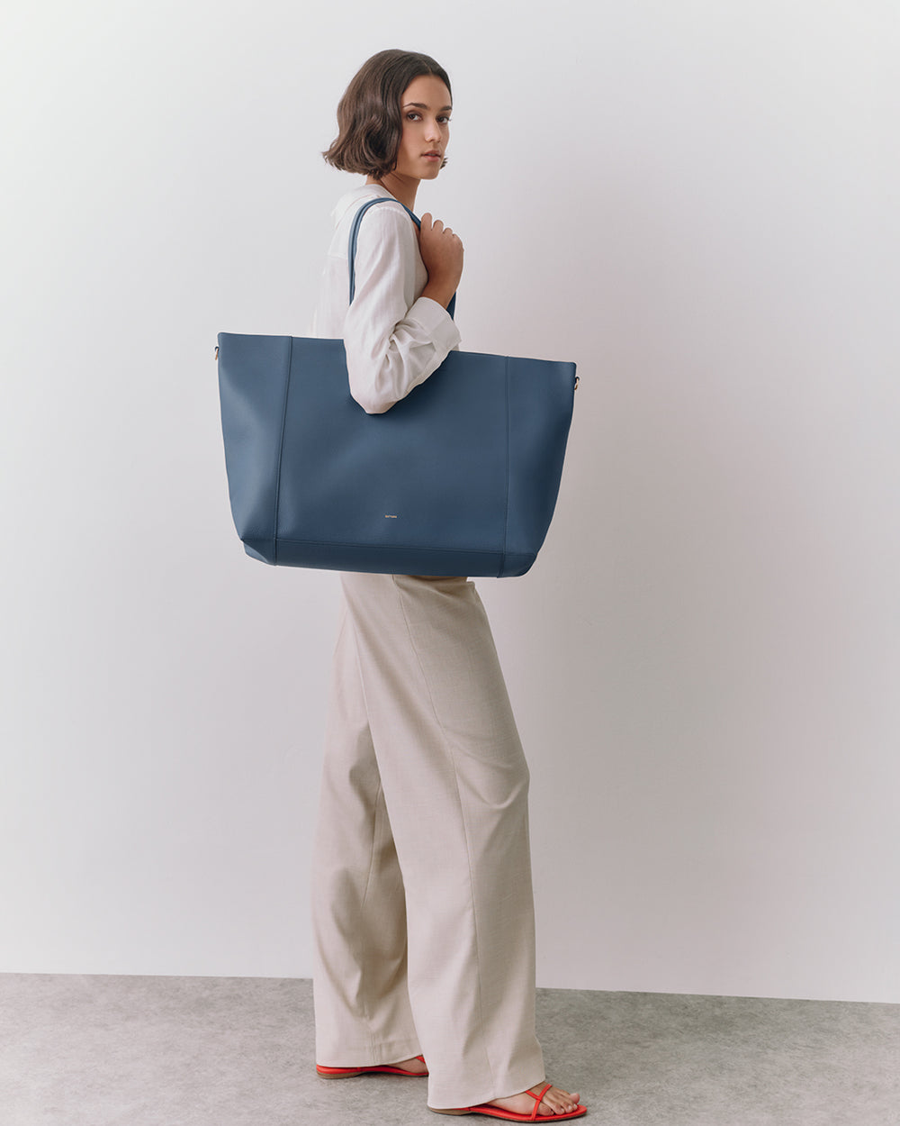 Person holding large tote bag wearing long-sleeve shirt and wide-leg pants.