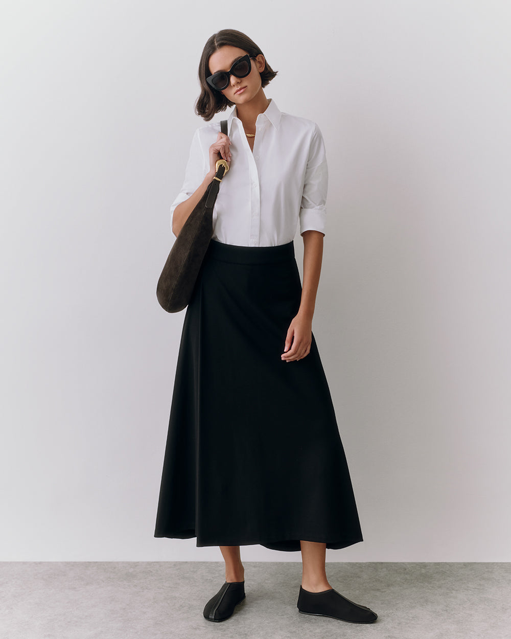 Person wears sunglasses and holds a bag, dressed in a button-up shirt and long skirt.