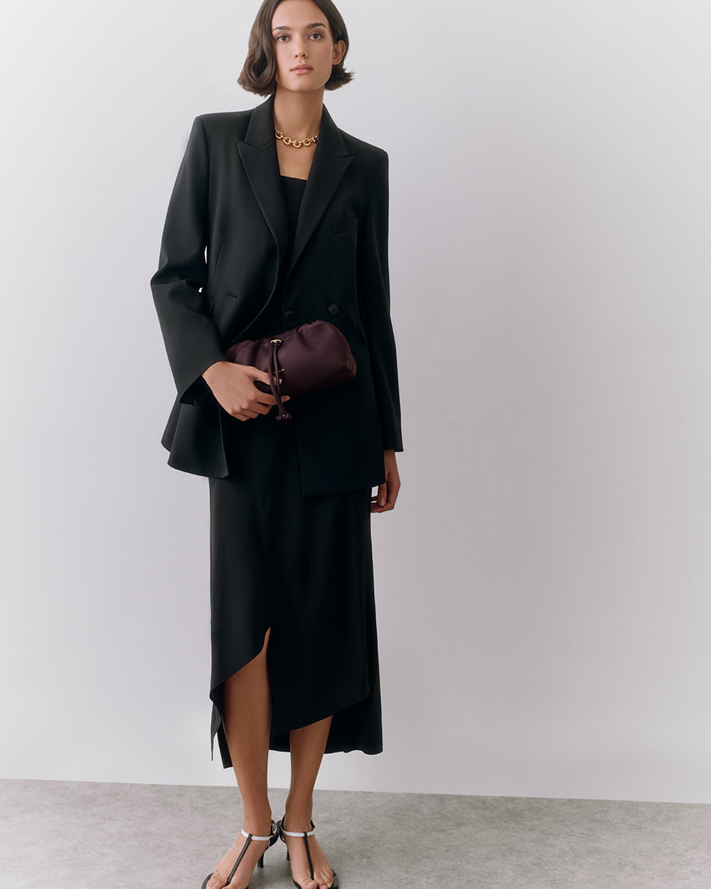 Person in jacket and dress holds a small bag, wearing sandals, standing against plain background.