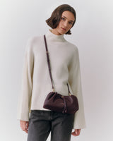 Person in sweater carrying a shoulder bag over their side standing against plain background.