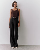 Person wearing a sleeveless top, wide-leg pants, and pointed shoes, standing against a wall.