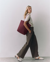 Person with large tote bag stands against a plain background, hands in pockets, wearing sandals.